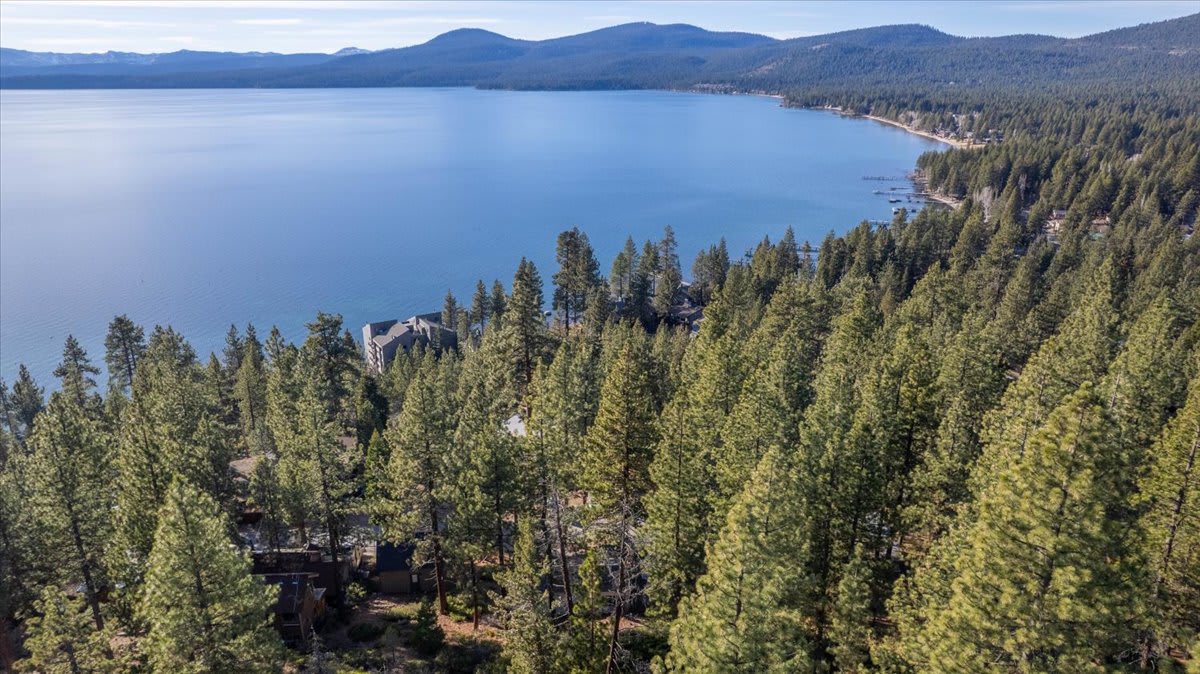 Aerial view of Kings Beach, showing a vacation rental amid tall pine trees near a serene lake and distant mountains.