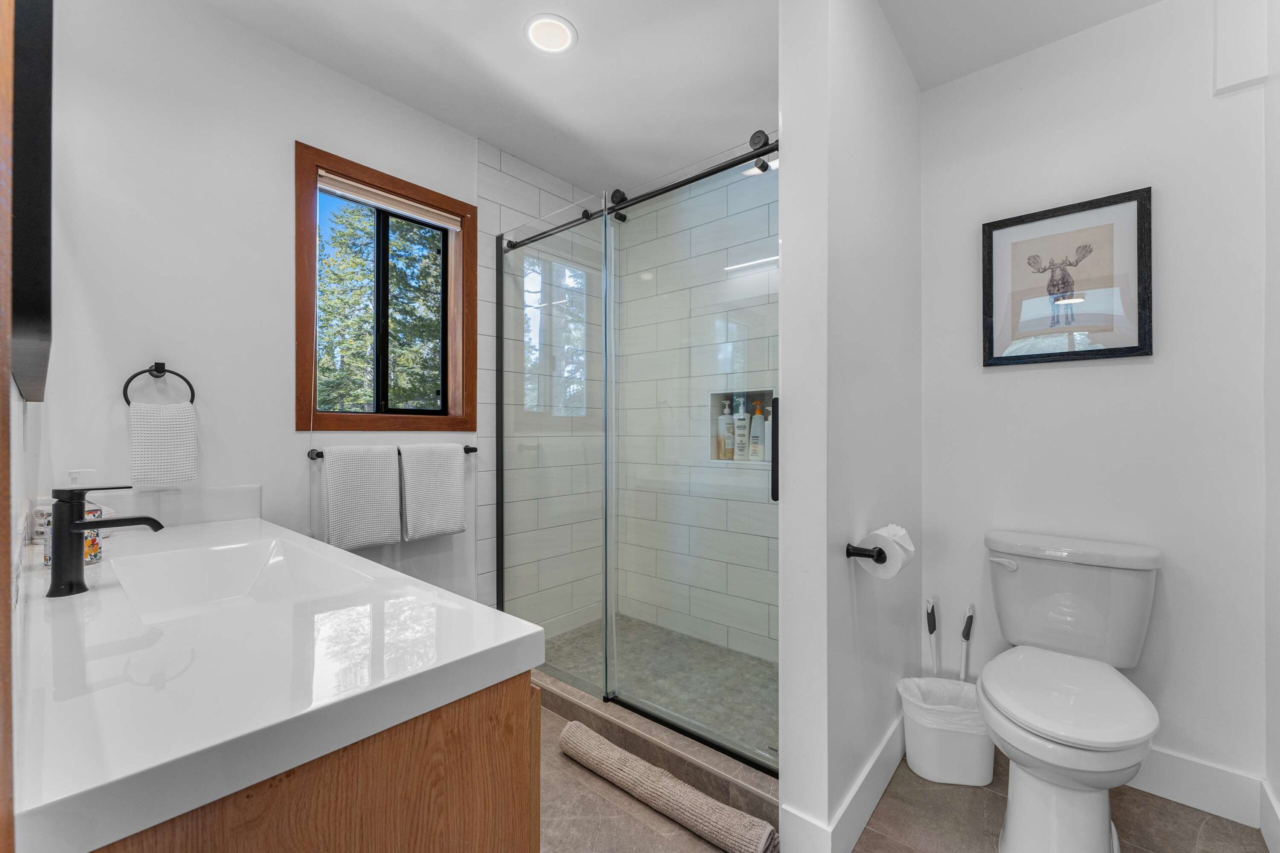Modern bathroom in Truckee vacation rental, featuring a glass shower, toilet, sink, and window with tree view.