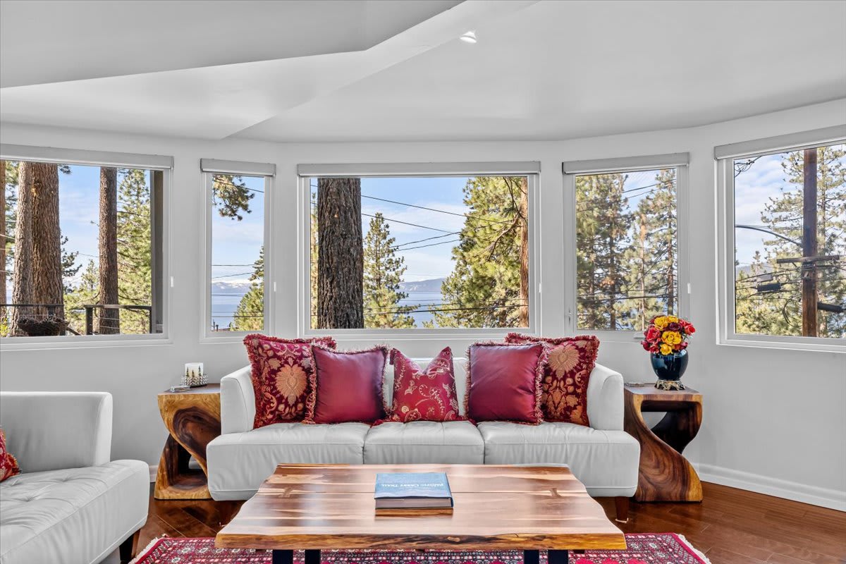 Bright living room in Kings Beach vacation rental with forest views through large windows, cozy white sofas, and red pillows.