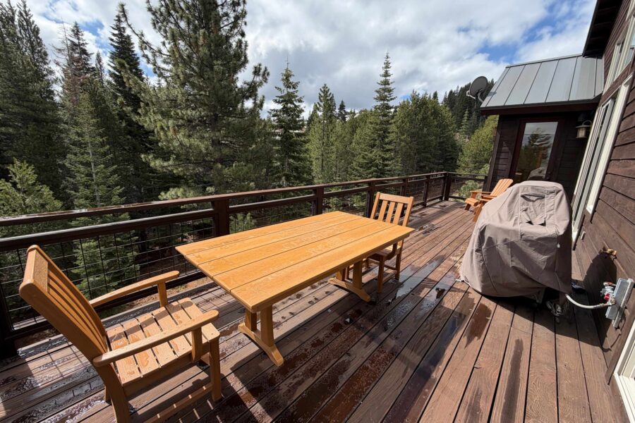Wooden deck of a Truckee vacation rental, featuring a dining table, chairs, and BBQ grill, surrounded by tall pine trees.