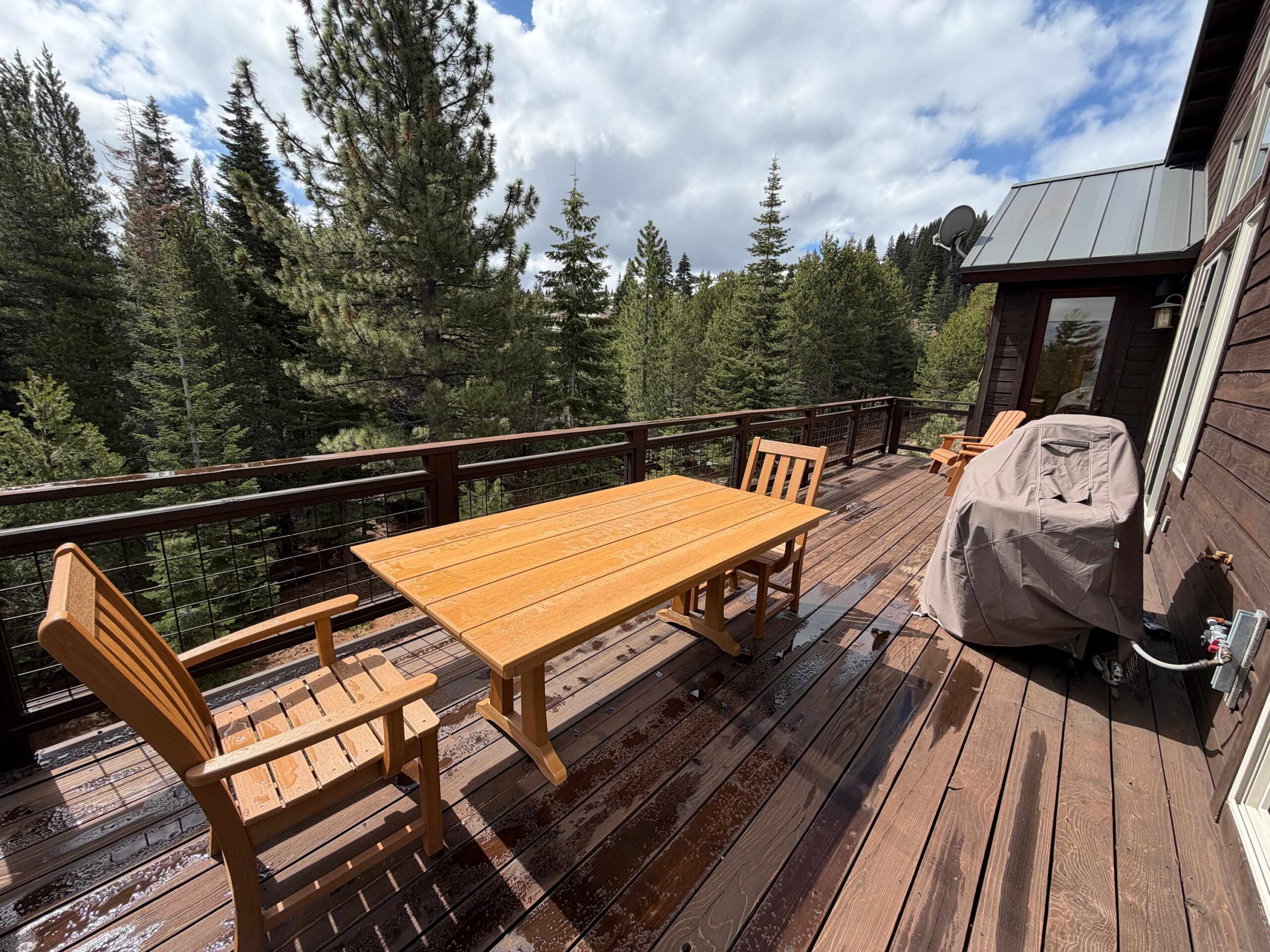 Wooden deck of a Truckee vacation rental, featuring a dining table, chairs, and BBQ grill, surrounded by tall pine trees.