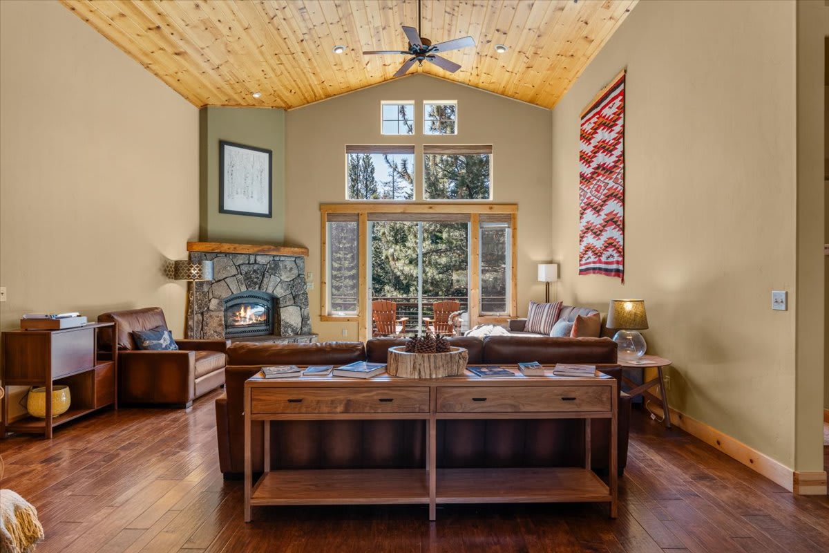 Cozy living room in a Truckee vacation rental with wooden ceiling, stone fireplace, and large windows overlooking trees.