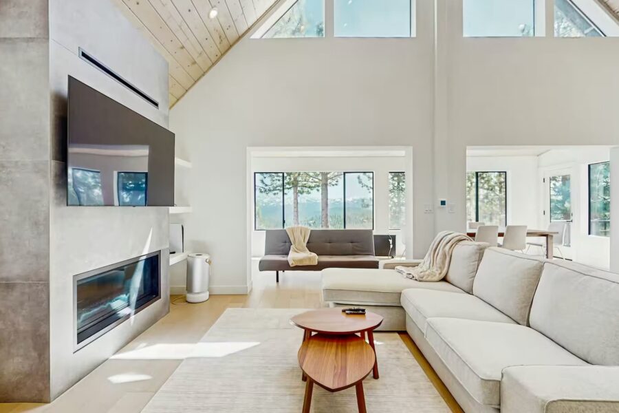 Modern living room with large windows in a Truckee vacation rental featuring a cozy sofa, TV, and fireplace.