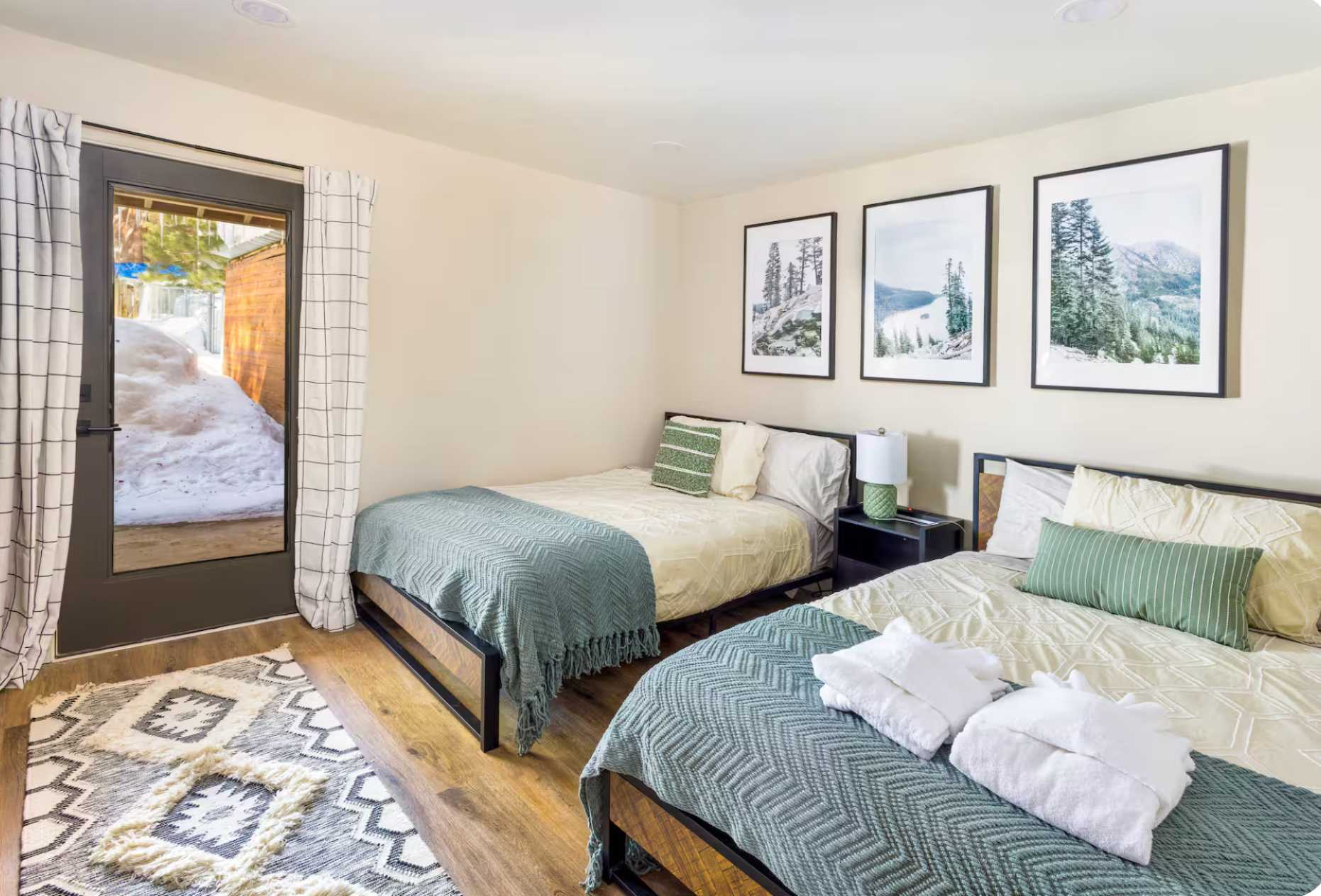 Two cozy beds in a Kings Beach vacation rental, decorated with nature art and a view of snow through the door.